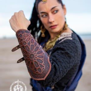 Puede incluir: Una protección de brazo de cuero marrón con intrincados diseños geométricos. La protección se lleva en el brazo izquierdo y tiene dos correas con hebillas.