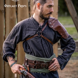 May include: A person wearing a dark grey shirt, leather shoulder armour, and a brown leather belt with a green sash. The person is holding a decorative pistol. The Outcast Props logo is visible in the upper left corner.