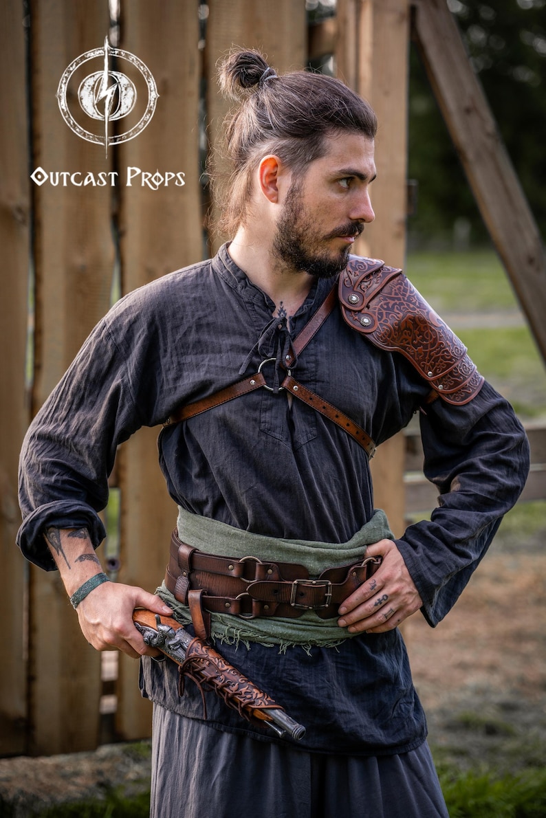 May include: A person wearing a dark grey shirt, leather shoulder armour, and a brown leather belt with a green sash. The person is holding a decorative pistol. The Outcast Props logo is visible in the upper left corner.