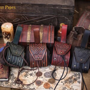Fourhandmade leather coin pouches displayed upright on a table, showing different color options including green, aged brown, red and black. Each pouch features detailed geometric embossing, brass hardware and a soft suede interior that expands for practical use. Designed to be worn on a belt, these pouches fit medieval, Viking, Celtic and dwarf aesthetics. Styled with candles, maps and wooden props, the image highlights their versatility for LARP, Renaissance fairs, cosplay, fantasy events