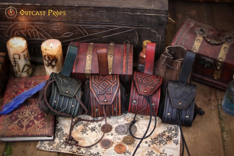 Fourhandmade leather coin pouches displayed upright on a table, showing different color options including green, aged brown, red and black. Each pouch features detailed geometric embossing, brass hardware and a soft suede interior that expands for practical use. Designed to be worn on a belt, these pouches fit medieval, Viking, Celtic and dwarf aesthetics. Styled with candles, maps and wooden props, the image highlights their versatility for LARP, Renaissance fairs, cosplay, fantasy events