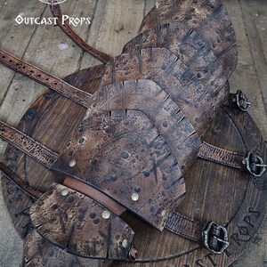 May include: A handcrafted, weathered-look leather and wood shield with riveted accents and buckle straps. The shield features the text "OUTCAST PROPS" and is designed for cosplay or display.