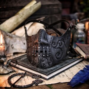 A black leather mask with intricate stitching and metal accents. The mask has a stylized face design with a prominent nose and mouth. The mask is lying on a table with a book and a feather pen. Death Wizard Leather Mask M2 - Handcrafted Mask for Dark mages witches and powerful sorcerers with Intricate Details and Metallic Finish