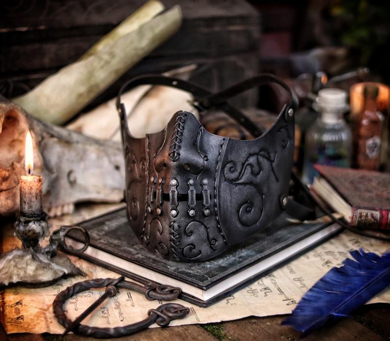 A black leather mask with intricate stitching and metal accents. The mask has a stylized face design with a prominent nose and mouth. The mask is lying on a table with a book and a feather pen. Death Wizard Leather Mask M2 - Handcrafted Mask for Dark mages witches and powerful sorcerers with Intricate Details and Metallic Finish