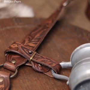 May include: Close-up of a brown leather strap with floral tooling and a metal buckle. A metal mug is attached to the strap. The text "Belt not included" is visible.
