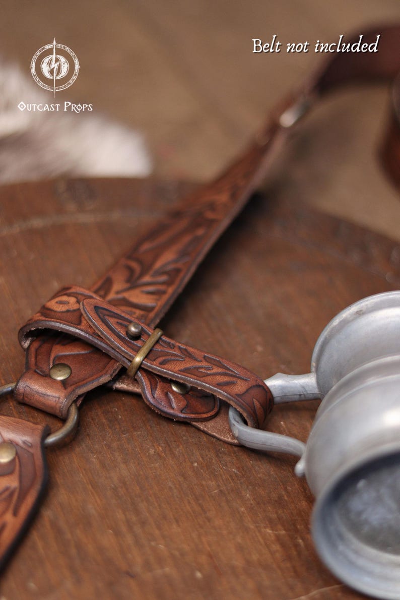 May include: Close-up of a brown leather strap with floral tooling and a metal buckle. A metal mug is attached to the strap. The text "Belt not included" is visible.
