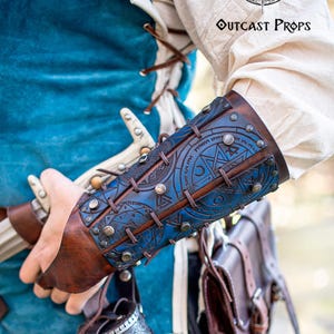 Detailed view of blue rune bracer worn by a fantasy adventurer with sword, scroll holder and belt accessories. Perfect LARP costume armor for alchemists, witch hunters, or steampunk characters. Handcrafted with engraved protection sigils, premium Spanish leather and epic weathered finish. Adjustable and practical for active cosplay or medieval reenactment.