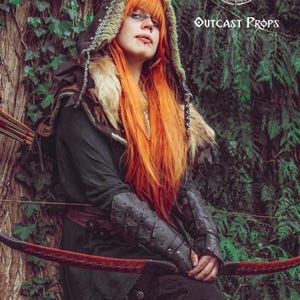 Female archer character with long orange hair and fur-trimmed hood leans against a tree, wearing black dragon scale bracers. The bracers add fierce elegance to LARP costumes, fantasy elf cosplay or rogue outfits. Made from genuine leather and aged by hand, these vambraces offer both comfort and bold style for medieval fairs or immersive fantasy adventures.