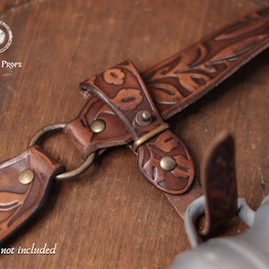 May include: Close-up of a brown leather prop with floral tooling and metal hardware. The leather strap features a decorative leaf pattern and is connected with brass-colored rivets and a metal ring. The text "Belt not included" is visible.