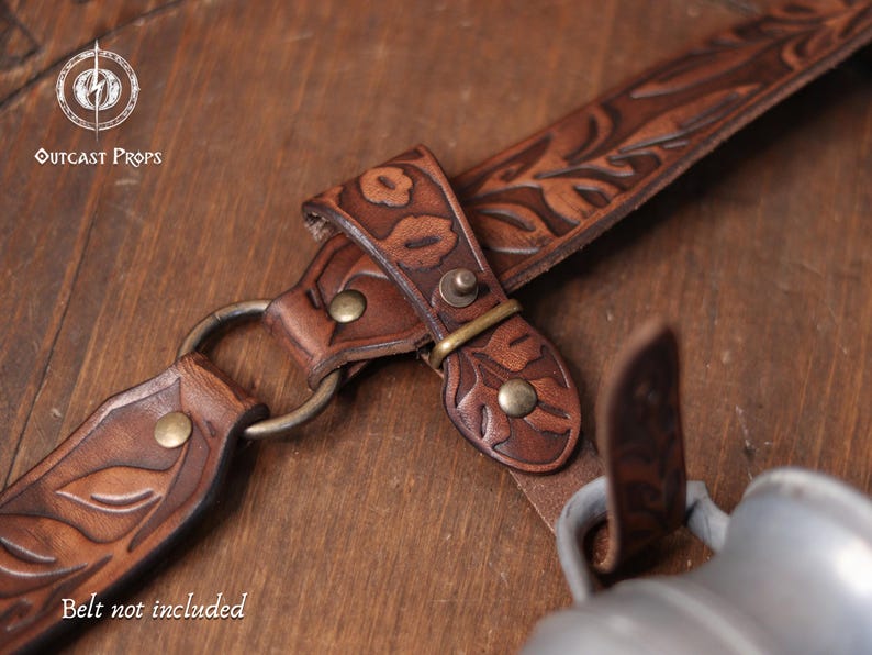 May include: Close-up of a brown leather prop with floral tooling and metal hardware. The leather strap features a decorative leaf pattern and is connected with brass-colored rivets and a metal ring. The text "Belt not included" is visible.