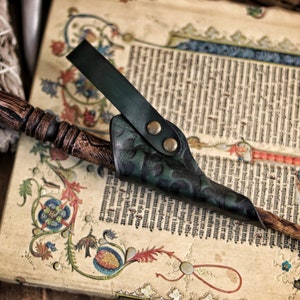 A green embossed leather wand holster holds a wooden wand on top of an illuminated medieval manuscript. The holster’s hand tooled details and riveted loop are clearly visible, with swirling patterns catching the light. The setting includes old books, sage bundles and rustic props, creating a magical atmosphere ideal for wizard cosplay, elven mages, witch costume accessories and renaissance faire wand holster use.