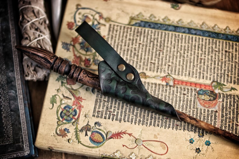 A green embossed leather wand holster holds a wooden wand on top of an illuminated medieval manuscript. The holster’s hand tooled details and riveted loop are clearly visible, with swirling patterns catching the light. The setting includes old books, sage bundles and rustic props, creating a magical atmosphere ideal for wizard cosplay, elven mages, witch costume accessories and renaissance faire wand holster use.