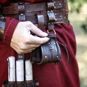 Detail view of a handcrafted leather coin pouch attached to a wide leather belt. Embossed geometric patterns inspired by Celtic and Viking motifs, aged brown leather finish, and drawstring closure. Compact pouch designed for coins, dice, keys, or small personal items. Ideal for fantasy characters, dwarven or medieval costumes, LARP, Renaissance fairs, or everyday leather accessories with character. Leather coin pouch with Celtic or Viking embossing,