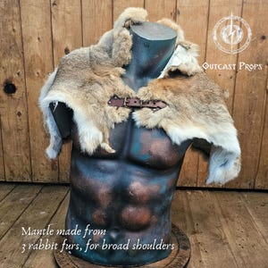 Pu&ograve; includere: Un mantello di pelliccia di coniglio con chiusura a fibbia in pelle marrone, esposto su un manichino. Il mantello &egrave; realizzato con tre pelli di coniglio e progettato per spalle larghe. Il testo "Outcast Props" &egrave; visibile.