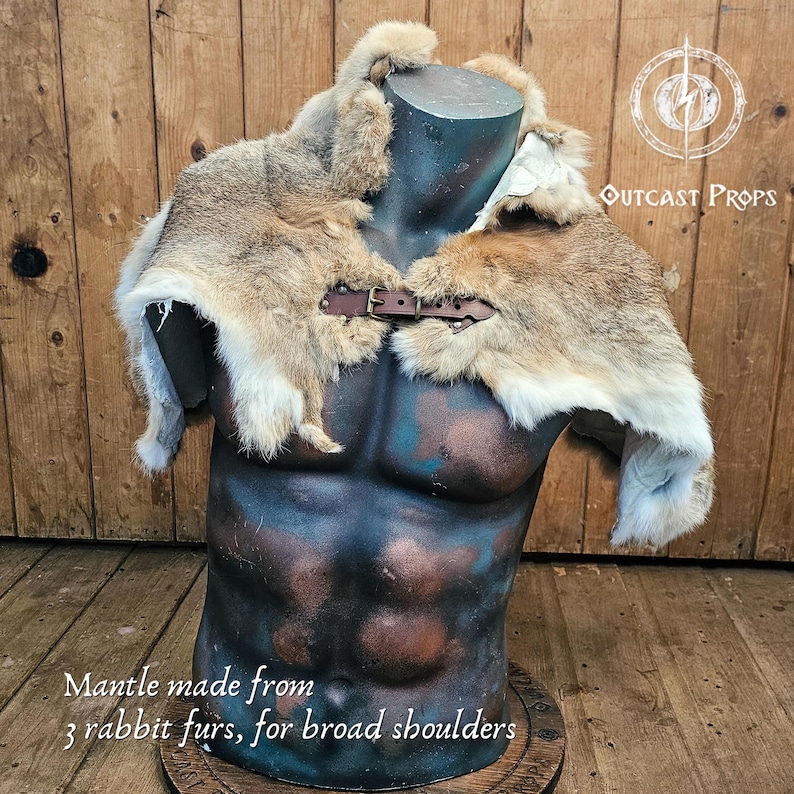 Pu&ograve; includere: Un mantello di pelliccia di coniglio con chiusura a fibbia in pelle marrone, esposto su un manichino. Il mantello &egrave; realizzato con tre pelli di coniglio e progettato per spalle larghe. Il testo "Outcast Props" &egrave; visibile.