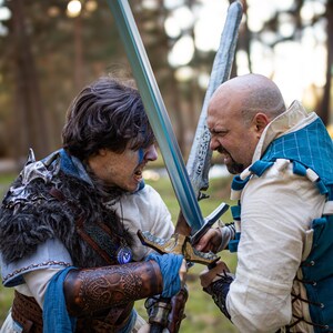 Dramatic forest battle scene between two fantasy warriors. The one with blue war paint wears brown leather bracers with Yggdrasil tree embossing, ideal for nordic warrior costume, viking leather bracers for larp, battle-ready cosplay armor, or medieval roleplay accessories. The dynamic action and intense expressions create a vivid moment of combat storytelling.