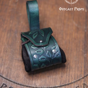 A green leather coin bag with an embossed floral design rests on a wooden surface with the Outcast Props logo above it. The pouch combines a structured vegetable tanned leather flap with soft black suede sides, creating an elegant elven accessory ideal for renfaire outfits, herbalist costumes, witches or nobles. The rounded shape and belt loop make it a practical leather coin bag or fantasy dice pouch for LARP and daily carry.