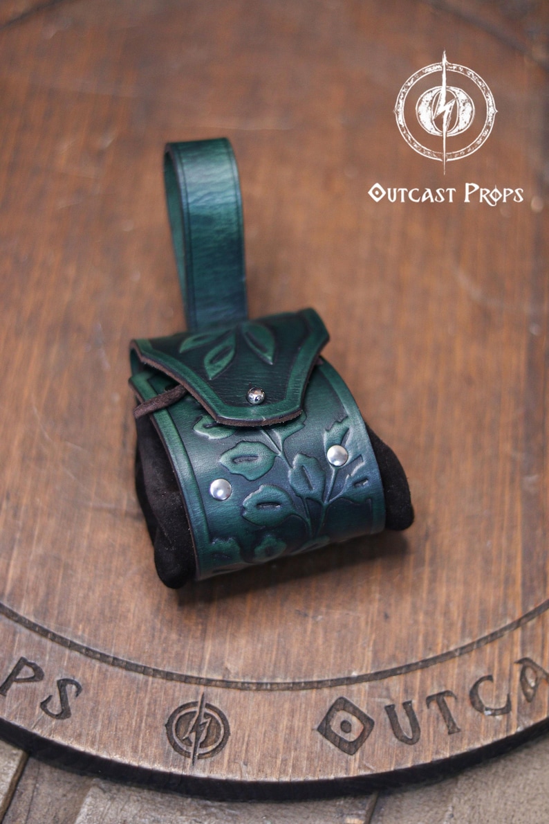 A green leather coin bag with an embossed floral design rests on a wooden surface with the Outcast Props logo above it. The pouch combines a structured vegetable tanned leather flap with soft black suede sides, creating an elegant elven accessory ideal for renfaire outfits, herbalist costumes, witches or nobles. The rounded shape and belt loop make it a practical leather coin bag or fantasy dice pouch for LARP and daily carry.