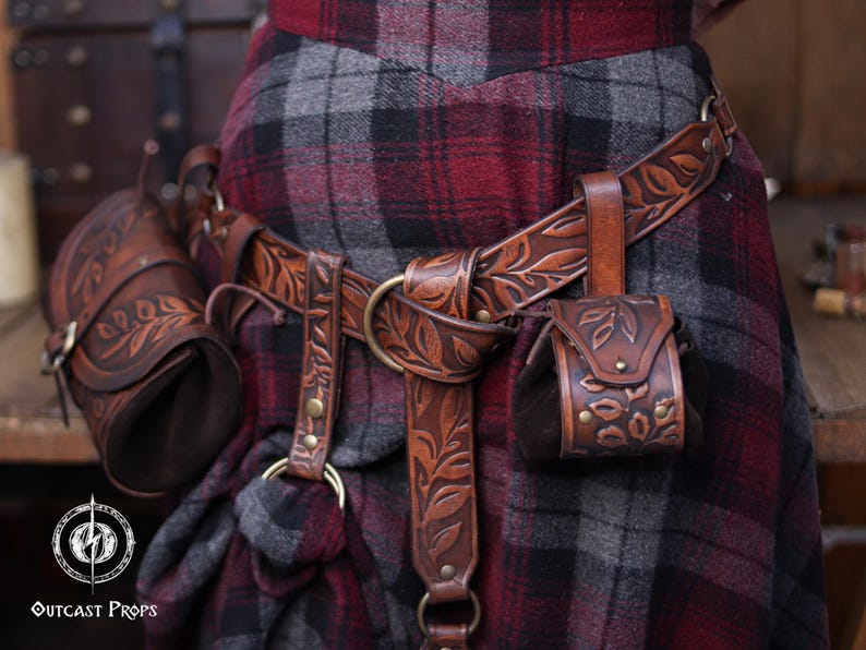 A full brown leather set with embossed botanical patterns is displayed on a person wearing a plaid skirt. The belt carries two accessories: a hipbag and a floral coin pouch. Brass rings and rivets complement the aged leather look. This renaissance faire inspired ensemble suits elves, witches, herbalists and nobles. The coin pouch works as a fantasy dice pouch or coin bag, offering practical storage for LARP and immersive costume wear.