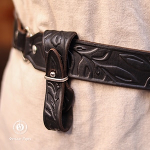 May include: Close-up of a black leather belt with embossed leaf patterns. The belt features a silver buckle and a matching strap. The design suggests a medieval or fantasy theme, suitable for costumes or themed outfits.