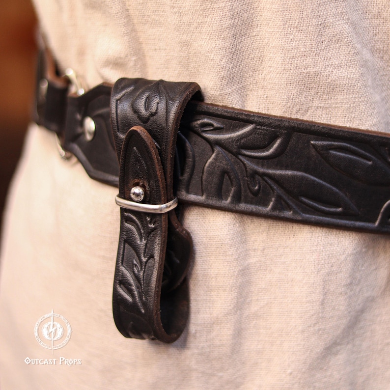 May include: Close-up of a black leather belt with embossed leaf patterns. The belt features a silver buckle and a matching strap. The design suggests a medieval or fantasy theme, suitable for costumes or themed outfits.