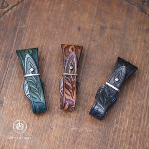 May include: Three leather straps with decorative leaf patterns and metal buckles. One is green, one is brown, and one is black. The straps are arranged on a wooden surface.