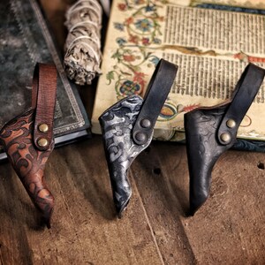 A row of three ornate leather wand holsters displayed on a wooden table beside an old illuminated manuscript and a spellbook. The holsters appear in aged brown, aged silver-black and matte black, each embossed with swirling patterns. Their belt loops are riveted with metal studs. Ideal for wizard cosplay, witch costume, larp wand holder needs, magic conventions and renaissance faire outfits. The warm rustic setting enhances their handcrafted texture and fantasy aesthetic.
