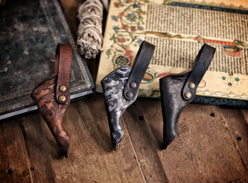 A row of three ornate leather wand holsters displayed on a wooden table beside an old illuminated manuscript and a spellbook. The holsters appear in aged brown, aged silver-black and matte black, each embossed with swirling patterns. Their belt loops are riveted with metal studs. Ideal for wizard cosplay, witch costume, larp wand holder needs, magic conventions and renaissance faire outfits. The warm rustic setting enhances their handcrafted texture and fantasy aesthetic.