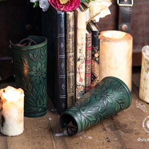 Two aged green floral leather bracers displayed among medieval style books and candles. The renaissance faire bracer design shows embossed leaves and flowers, highlighting artisan craftsmanship and natural shading. Ideal as an elf cosplay accessory, herbalist gear or fae costume accessory. This floral arm guard set fits renaissance faire costumes, larp bracer looks and romantic fantasy characters. Fae costume, ren faire, druid larp, leaf design, floral design.