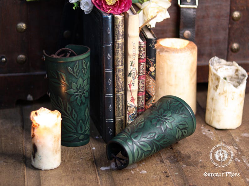 Two aged green floral leather bracers displayed among medieval style books and candles. The renaissance faire bracer design shows embossed leaves and flowers, highlighting artisan craftsmanship and natural shading. Ideal as an elf cosplay accessory, herbalist gear or fae costume accessory. This floral arm guard set fits renaissance faire costumes, larp bracer looks and romantic fantasy characters. Fae costume, ren faire, druid larp, leaf design, floral design.