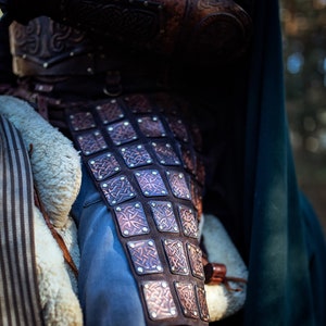 May include: Close-up of a brown leather armor with intricate Celtic knot designs. The armor is worn over blue jeans and has a row of metal rivets.