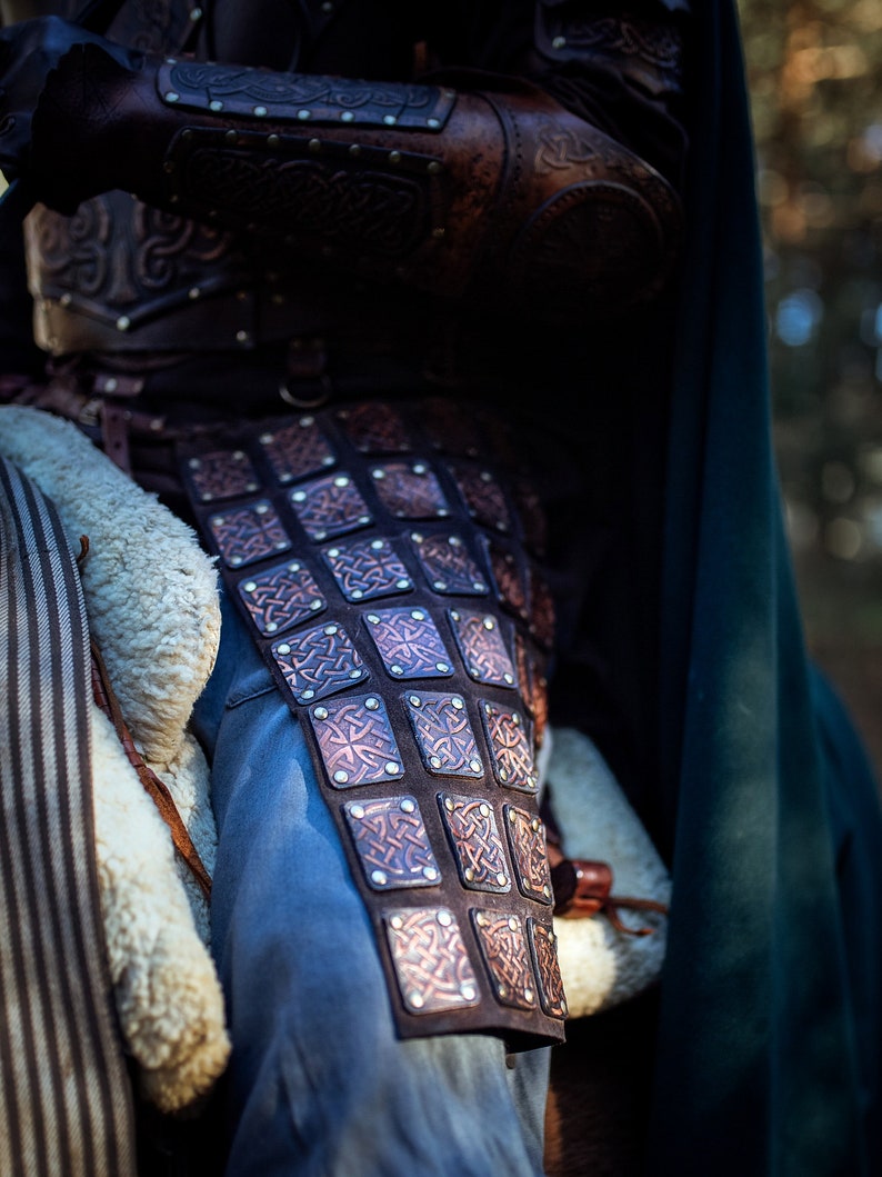 May include: Close-up of a brown leather armor with intricate Celtic knot designs. The armor is worn over blue jeans and has a row of metal rivets.