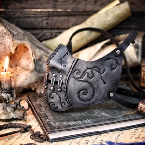 A black leather mask with intricate silver detailing and a floral design. The mask has a strap that goes around the head and is secured with a metal buckle. Death Wizard Leather Mask M2 - Handcrafted Mask for Dark mages witches and powerful sorcerers with Intricate Details and Metallic Finish