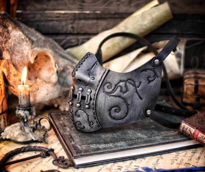 A black leather mask with intricate silver detailing and a floral design. The mask has a strap that goes around the head and is secured with a metal buckle. Death Wizard Leather Mask M2 - Handcrafted Mask for Dark mages witches and powerful sorcerers with Intricate Details and Metallic Finish