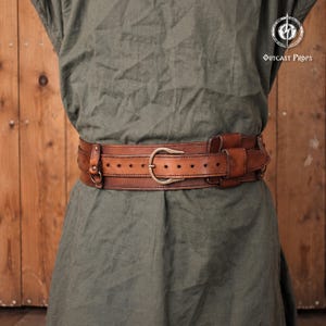 May include: A wide brown leather belt with a double-layered design, featuring a central buckle, multiple adjustment holes, and integrated metal rings. The belt is worn over a plain olive green tunic. The text OUTCAST PROPS is visible.