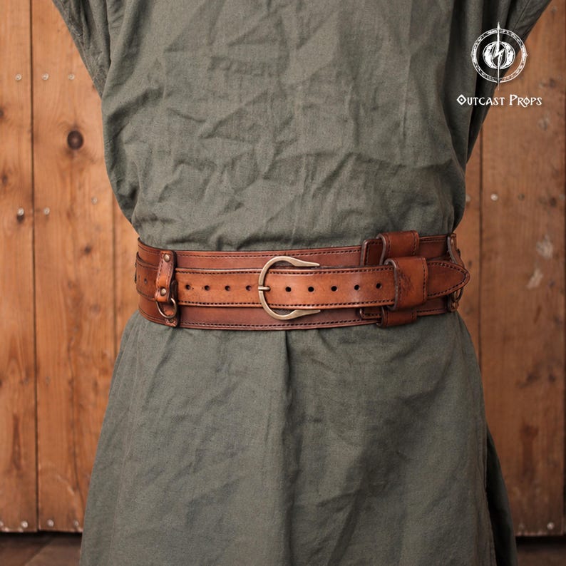May include: A wide brown leather belt with a double-layered design, featuring a central buckle, multiple adjustment holes, and integrated metal rings. The belt is worn over a plain olive green tunic. The text OUTCAST PROPS is visible.