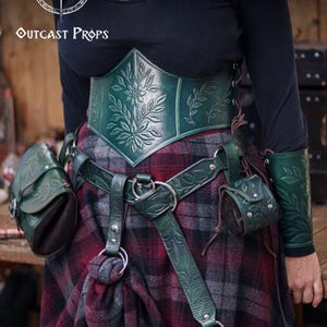 A model wears a full green leather set with embossed floral patterns, including corset, belt, hipbag and coin pouch. The pieces have botanical carvings and soft suede bodies, creating an elegant elven or herbalist look. The plaid skirt enhances the renaissance faire style. This fantasy outfit shows how the floral coin pouch attaches to the belt, making it suitable for witches, nobles and renfaire characters who need a practical accessory for coins or dice.