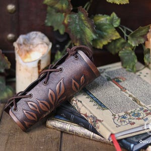 Aged brown floral leather bracer displayed horizontally over illuminated medieval style pages with candles around it. The renaissance faire bracer shows detailed embossed leaves that form a refined floral arm guard suited to elf cosplay accessory, herbalist gear or fae costume accessory use. Warm light enhances its artisan finish, making it an elegant larp bracer for renaissance faire and fantasy outfits. Fae costume, ren faire, druid larp, leaf design, floral design.