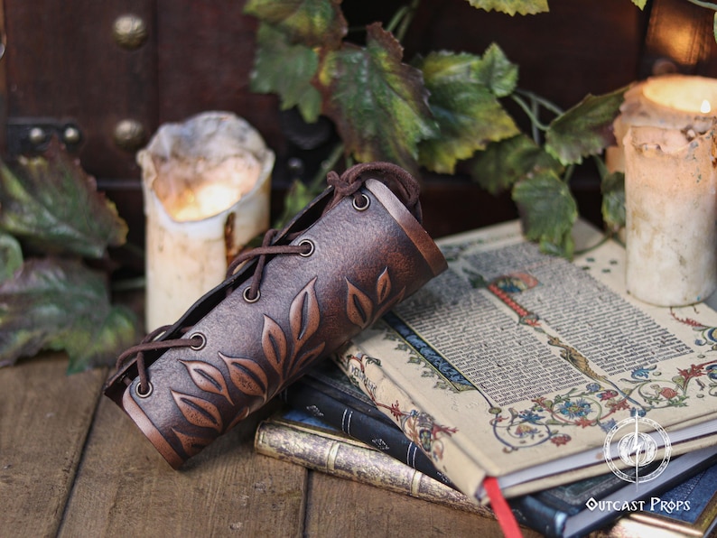 Aged brown floral leather bracer displayed horizontally over illuminated medieval style pages with candles around it. The renaissance faire bracer shows detailed embossed leaves that form a refined floral arm guard suited to elf cosplay accessory, herbalist gear or fae costume accessory use. Warm light enhances its artisan finish, making it an elegant larp bracer for renaissance faire and fantasy outfits. Fae costume, ren faire, druid larp, leaf design, floral design.