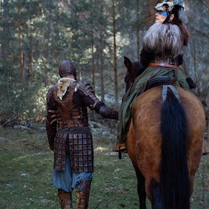 May include: A person wearing leather armor and a helmet stands in a forest, looking at a person riding a brown horse. The person on the horse is wearing a horned headdress and a leather vest.