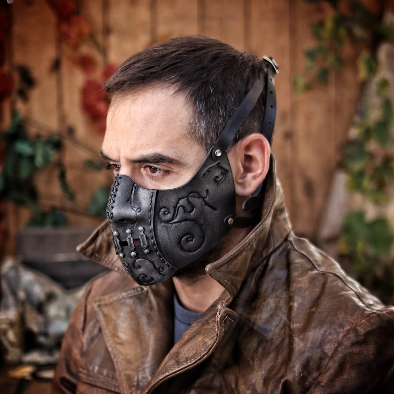 A black leather face mask with intricate metal accents and a floral design. The mask has a strap that goes over the head and is secured with a buckle. Death Wizard Leather Mask M2 - Handcrafted Mask for Dark mages witches and powerful sorcerers with Intricate Details and Metallic Finish