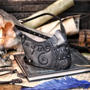 A black leather mask with intricate silver detailing and a strap. The mask has a pointed nose and a curved mouth. The mask is lying on a book with a blue feather next to it. Death Wizard Leather Mask M2 - Handcrafted Mask for Dark mages witches and powerful sorcerers with Intricate Details and Metallic Finish