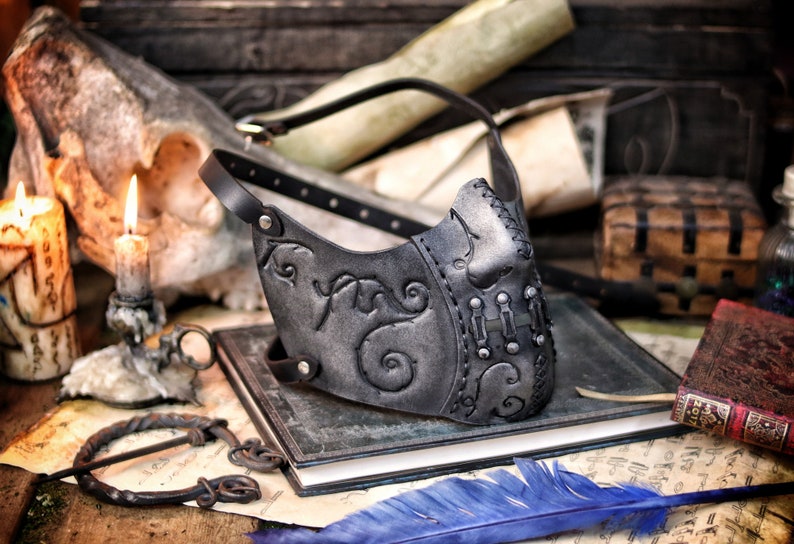A black leather mask with intricate silver detailing and a strap. The mask has a pointed nose and a curved mouth. The mask is lying on a book with a blue feather next to it. Death Wizard Leather Mask M2 - Handcrafted Mask for Dark mages witches and powerful sorcerers with Intricate Details and Metallic Finish