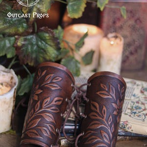 Two aged brown floral leather bracers with laced fastening lay on antique books surrounded by ivy and candlelight. The renaissance faire bracer design highlights natural shading and embossed leaves, creating a handcrafted floral arm guard ideal as an elf cosplay accessory or fae costume accessory. The rustic scene enhances their larp bracer appeal for herbalists, forest characters and medieval fantasy costumes. Fae costume, ren faire, druid larp, leaf design, floral design.