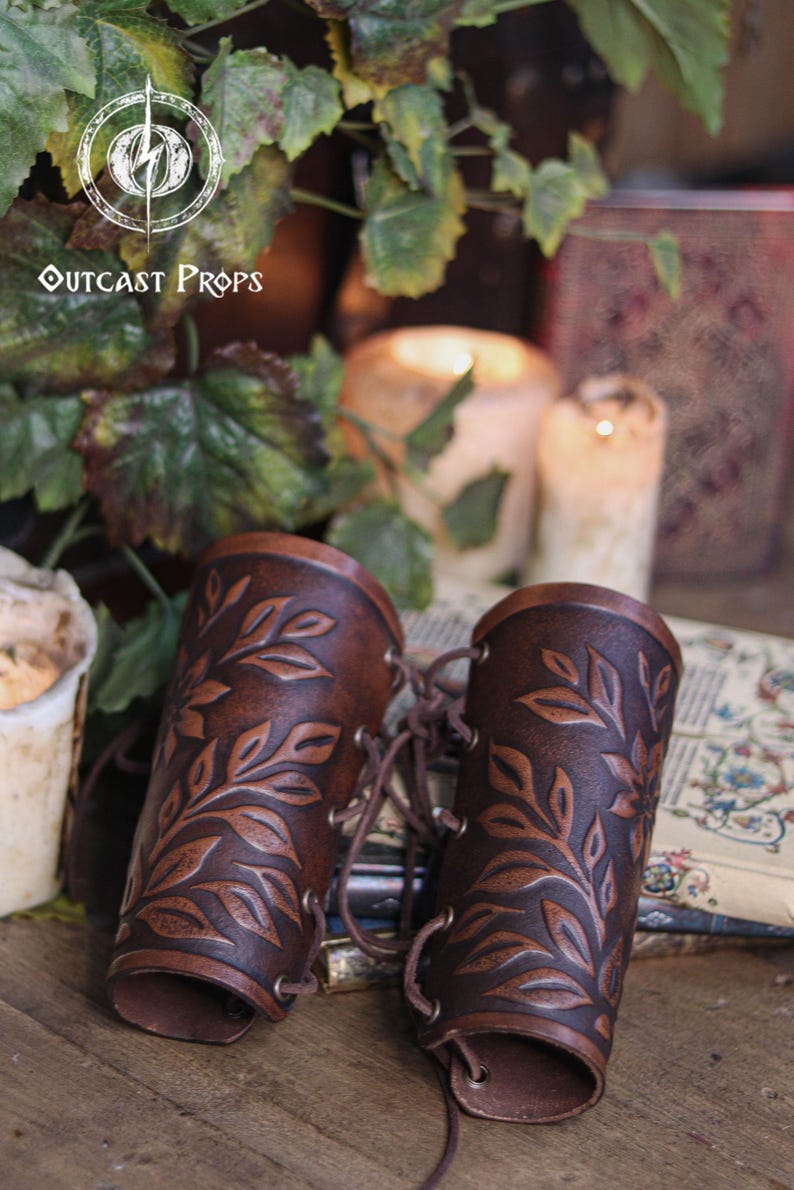 Two aged brown floral leather bracers with laced fastening lay on antique books surrounded by ivy and candlelight. The renaissance faire bracer design highlights natural shading and embossed leaves, creating a handcrafted floral arm guard ideal as an elf cosplay accessory or fae costume accessory. The rustic scene enhances their larp bracer appeal for herbalists, forest characters and medieval fantasy costumes. Fae costume, ren faire, druid larp, leaf design, floral design.
