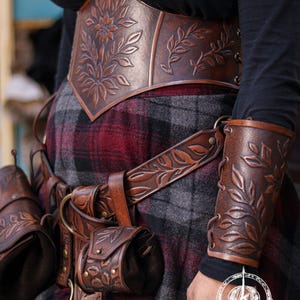 Close up of a brown floral leather coin pouch being attached to a carved leather belt, perfect for Renaissance Faire elves, herbalists or witches. The vegetable tanned leather shows embossed leaves and a soft suede inner bag. This LARP friendly pouch fits belts up to 6 cm and expands to carry coins or small items, maintaining a refined renaissance noble look. The image helps viewers understand how the loop attaches securely.