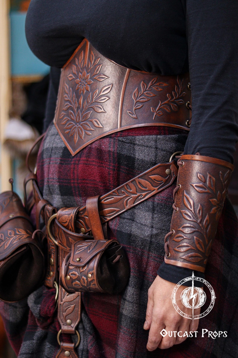 Close up of a brown floral leather coin pouch being attached to a carved leather belt, perfect for Renaissance Faire elves, herbalists or witches. The vegetable tanned leather shows embossed leaves and a soft suede inner bag. This LARP friendly pouch fits belts up to 6 cm and expands to carry coins or small items, maintaining a refined renaissance noble look. The image helps viewers understand how the loop attaches securely.