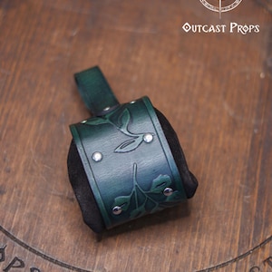 Close up of the back of a green embossed leather coin bag displayed on a round wooden board engraved with the Outcast Props logo. The pouch features botanical leaf motifs, silver rivets and soft suede sides. Designed as an elven herbalist bag, it attaches to the belt and works perfectly for carrying coins, keys or dice at a renaissance faire, renfaire or LARP event. Its handcrafted look and floral pattern make it a refined fantasy dice pouch.
