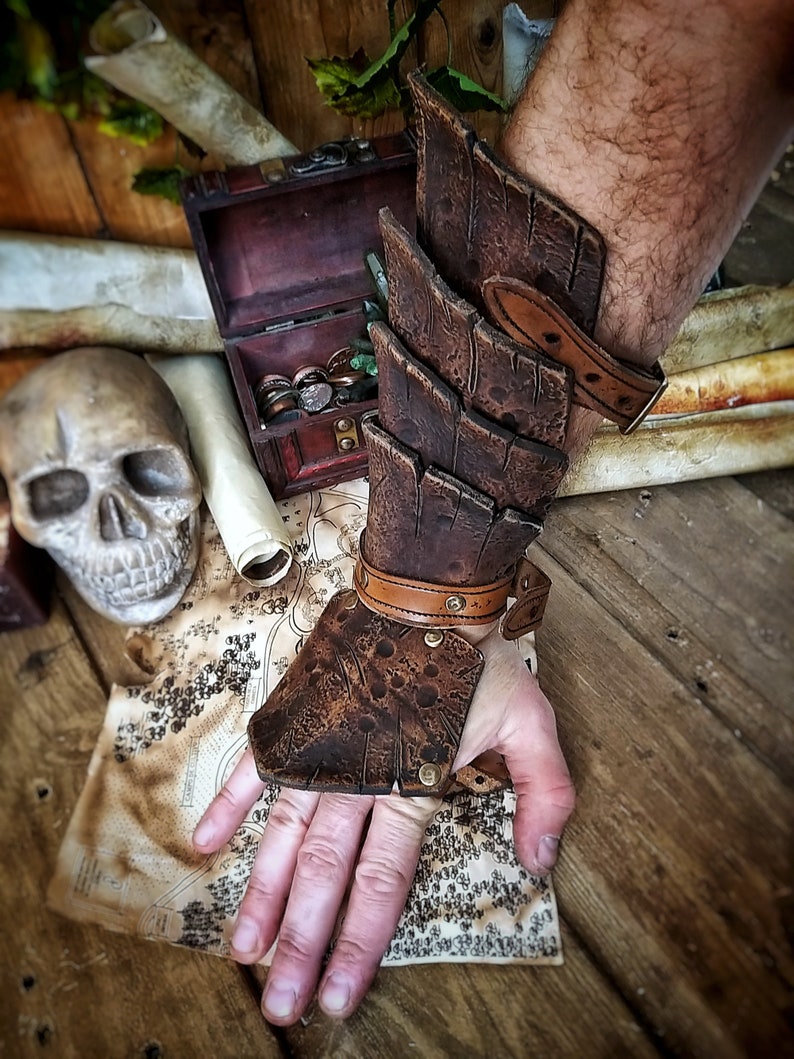 Close-up of a brown leather dragon scale bracer worn on a male hand, displayed over a vintage map with coins, a skull, and scrolls. This handmade bracer is ideal for LARP, pirate cosplay, monster hunters, or fantasy warriors. Crafted from premium vegetable-tanned leather and finished with Outcast Props' signature aging techniques, each piece is unique and tells a story straight from an epic adventure.