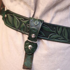 May include: A dark green leather belt with a leaf pattern, secured with a silver buckle. The belt is worn over a light-colored garment. The text "Belt not included" is visible. The Outcast Props logo is in the bottom right corner.